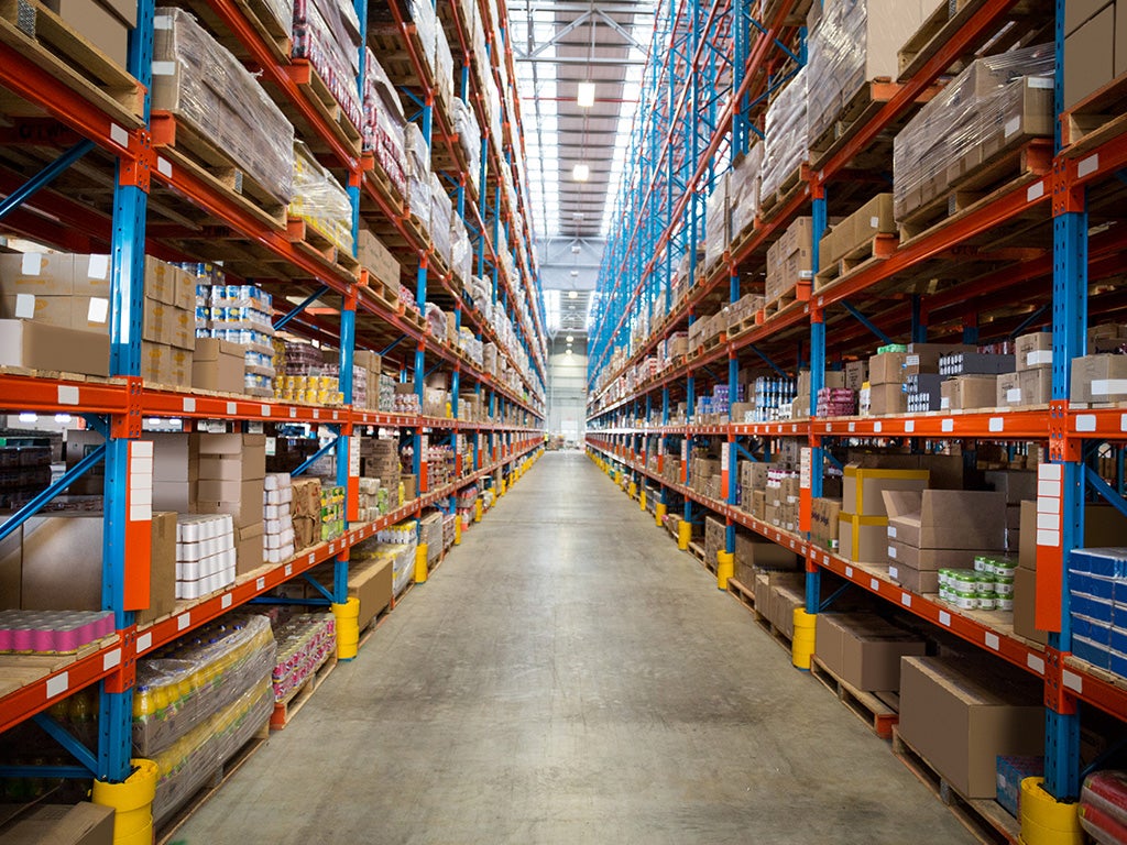 A wide aisle in a large warehouse with tall shelves on both sides filled with various boxed goods. The shelves are supported by blue and orange metal beams, and the floor is made of concrete. The warehouse is well-lit with overhead lighting.
