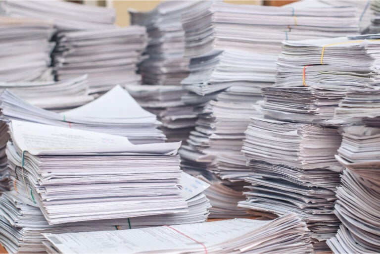 Stacks of numerous paper documents piled high on a desk, organized with rubber bands.