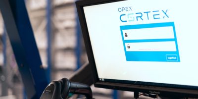 Computer monitor displaying a login screen with the text "OPEX CORTEX" next to a barcode scanner in an industrial setting, seamlessly integrated into an Infinity® AS/RS by OPEX®.